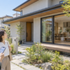 住宅情報をスマートフォンで調べながら、実際の戸建て住宅の外観や窓、庭とのつながりを確認するイメージ